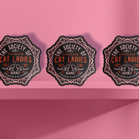 Round sticker with ornate border showing two cats and text "The Society of Childless Cat Ladies Against Couch Fuckers" in pink and purple versions on black background.